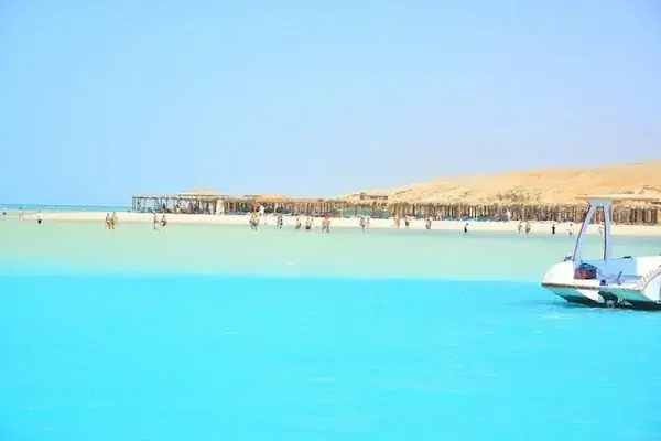 Wide turquoise lagoon with people wading near a sandy shoreline lined with straw huts, a speedboat approaching in the foreground