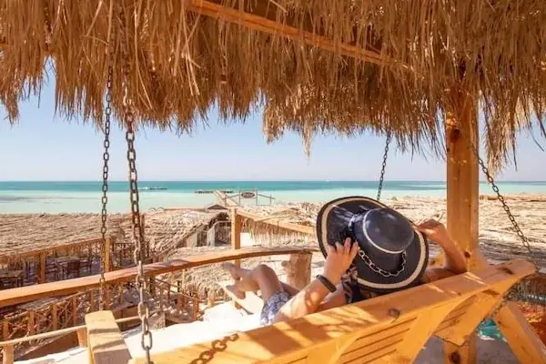 Person in a striped hat relaxing on a wooden swing bench under a straw roof, looking out at the turquoise sea