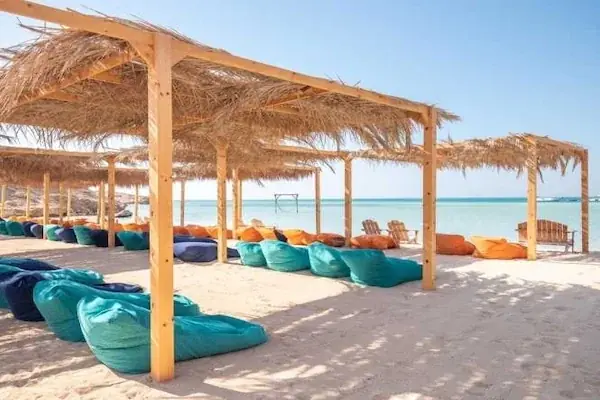 Row of straw-roofed cabanas with teal, navy, and orange bean bags arranged on white sand facing the sea