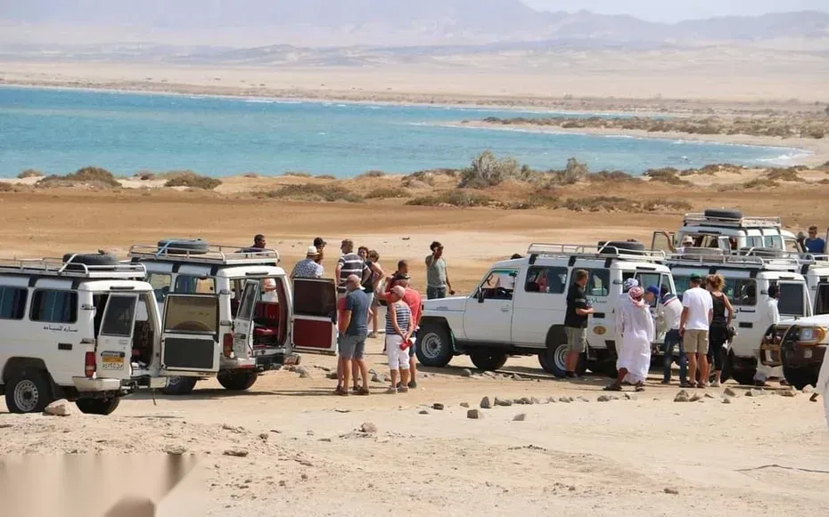 Line of parked safari jeeps and tourists gathered on a bluff overlooking a turquoise coastal lagoon