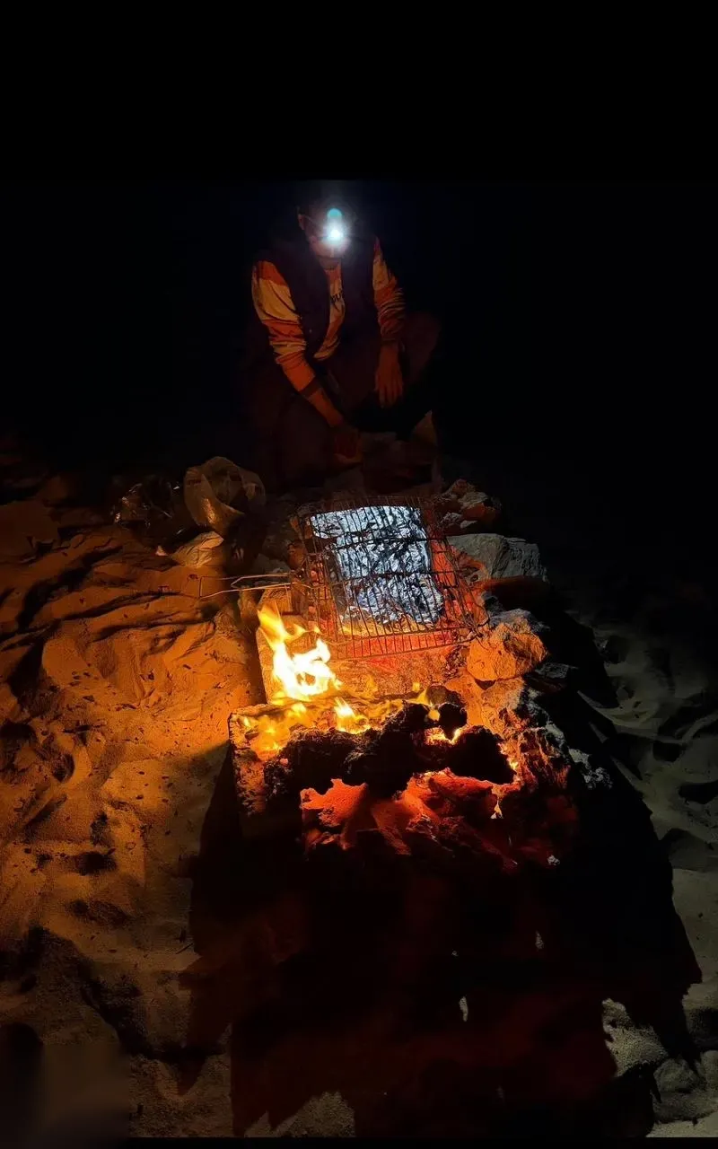 Person wearing a headlamp grilling meat on a wire rack over glowing campfire coals at night in the desert