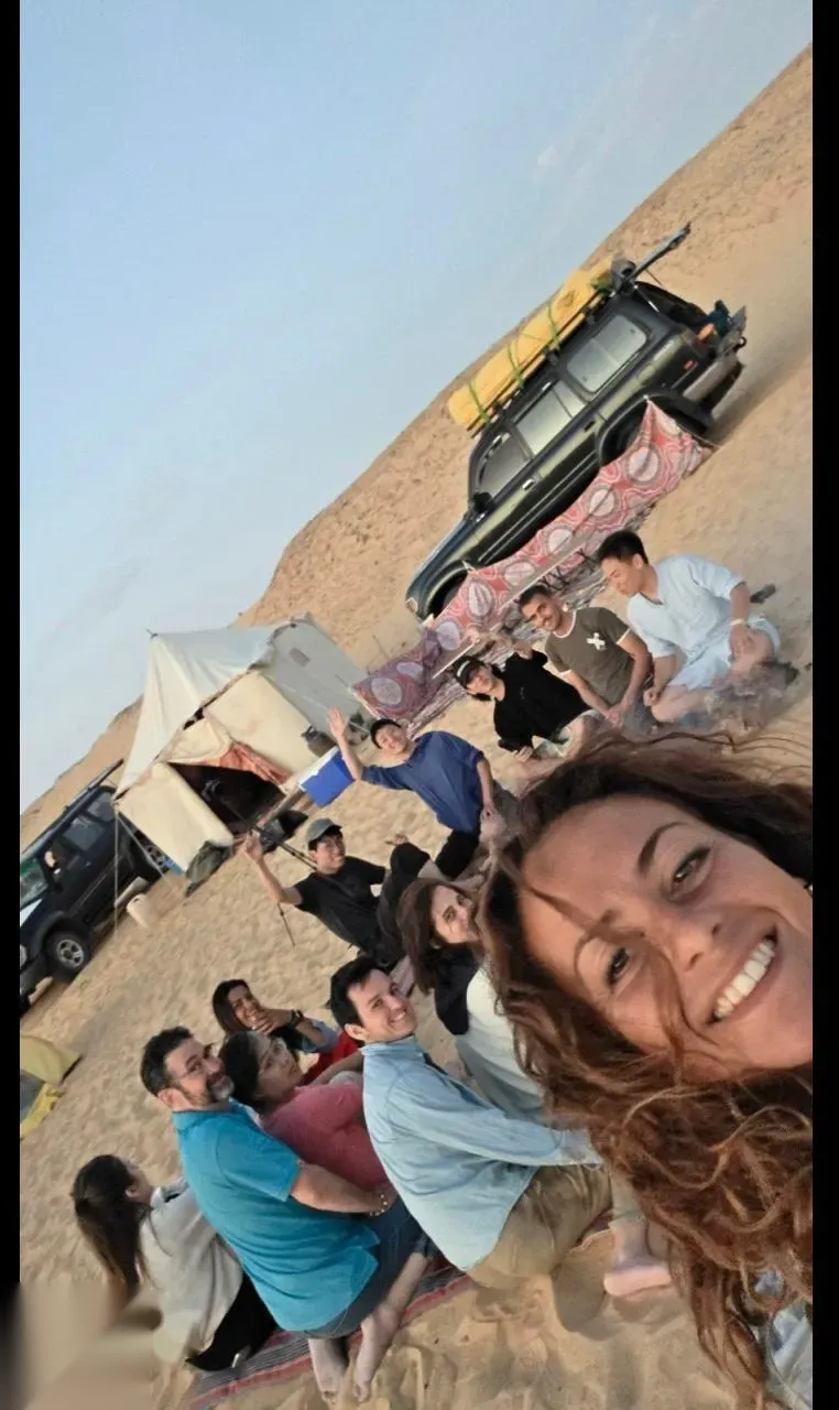 Tilted group selfie of safari travelers sitting on rugs near tents and a black 4x4 with sand dunes at sunset