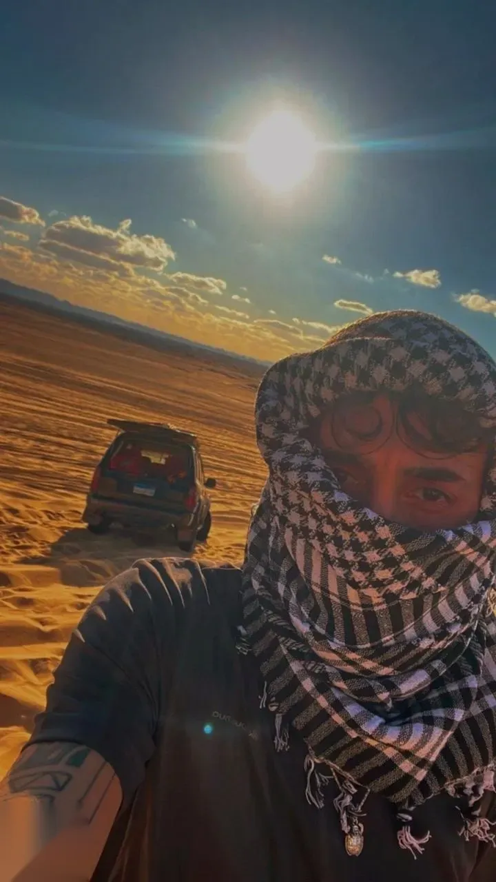 Man wrapped in a checkered keffiyeh scarf taking a selfie in front of a black 4x4 in the desert with the sun overhead