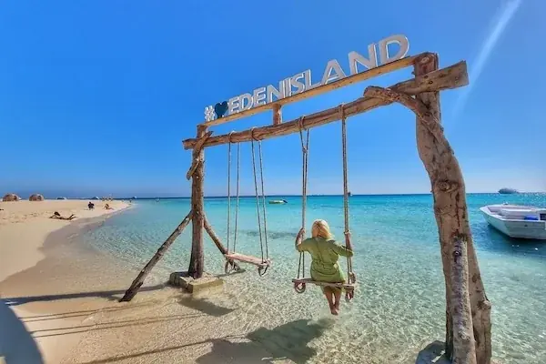 Beach lined with thatched palm-frond huts and loungers beside a turquoise lagoon, with wooden platforms partly submerged in the shallow water